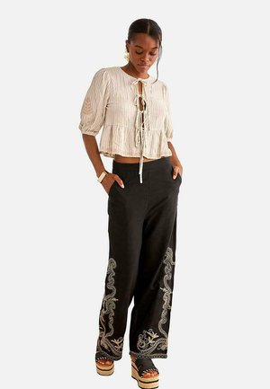 Mujer con blusa beige corta con lazo delantero y mangas abullonadas, pantalones negros de pierna ancha con bordado blanco y sandalias de plataforma.