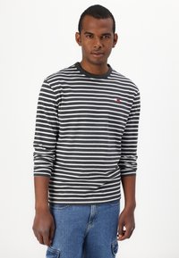 Rutet langt erme skjorte i mørkegrå og hvit med rund hals. Har en liten rød og blå logo på brystet. Kombinert med denimshorts.