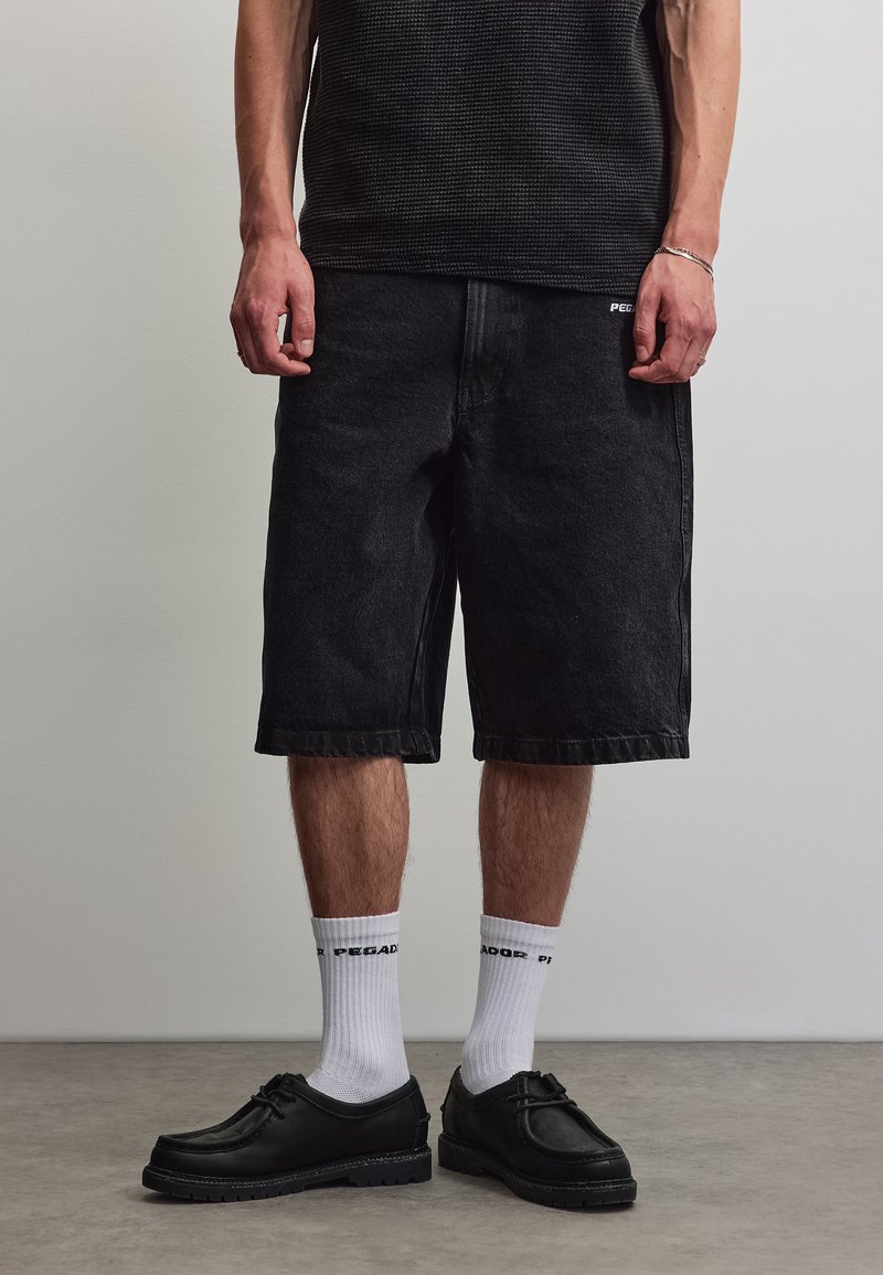 Mann trägt schwarze knielange Shorts, weiße Crew-Socken mit Text, schwarze Schnürschuhe und ein schwarzes strukturiertes Kurzarmhemd.