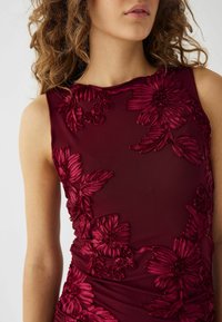 Burgundy klänning med blommig broderi och upphöjda blad, ärmlös design och textil drapering vid midjan.