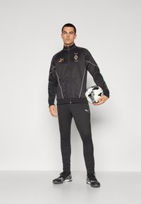 Survêtement de sport noir avec des accents blancs, comprenant une veste zippée, un pantalon ajusté et des chaussures de sport synthétiques noires. Tient un ballon de football.
