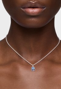 Srebrny naszyjnik z łańcuszkiem, z zawieszką w kształcie niedźwiedzia w odcieniach niebieskiego i ciemnoniebieskiego, spoczywający na skórze o gładkiej teksturze.