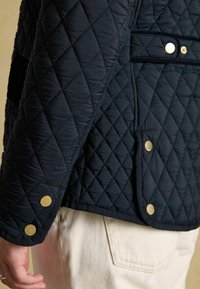 Jachetă matlasată bleumarin cu un material texturat, butoane din metal aurit și margini contrastante. Prim-plan al detalii de pe manchetă.