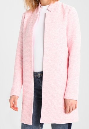 Femme portant un manteau ouvert texturé rose clair sur un haut blanc et un jean bleu devant un fond uni.