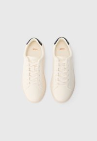 Pāris gaiši krēmkrāsas mežģīņu sporta apavu ar melnām papēža cilpām, ar uzrakstu "BOSS" iekšpusē, attēloti uz gaiši pelēka fona.