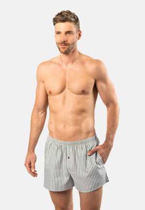 Man with short light brown hair, beard, and muscular build wearing striped boxer shorts, standing with one hand in pocket.