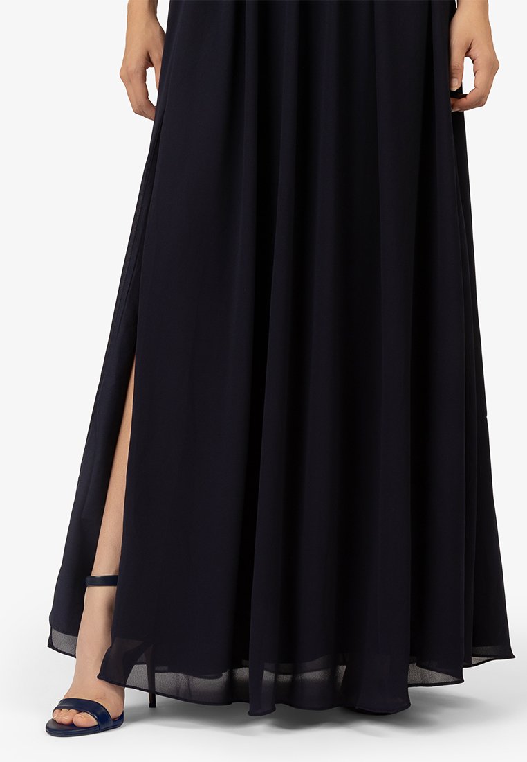 Granatowa sukienka maxi z lekkiego materiału z bocznymi rozcięciami, zestawiona z ciemnoniebieskimi sandałami na paski. Gładka tekstura, falujący krój.
