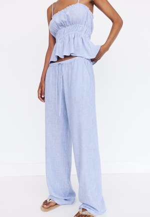 Mujer con pantalones anchos a rayas de color azul claro y sueltos, y un top sin mangas fruncido a juego, con sandalias, de pie frente a un fondo blanco.