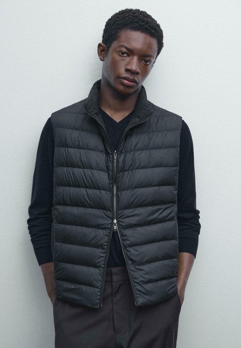 Jeune homme aux cheveux courts et bouclés, portant un gilet matelassé noir sur une chemise à manches longues noire, se tenant détendu avec les mains dans les poches contre un mur clair.