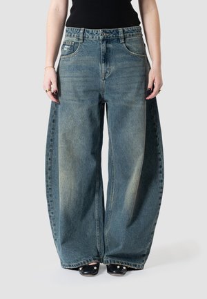 Personne portant un jean bleu délavé à jambes larges avec un haut noir et des chaussures noires ornées de détails en perles, posant devant un fond uni.