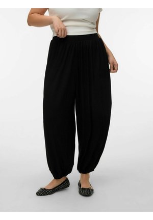 Kvinne iført løstsittende svarte bukser med elastisk midje og rynkede mansjetter, og svarte og hvite mønstrede flate sko, stående foran en ensfarget bakgrunn.