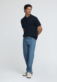 Svart pikétröja med kort ärm och en diskret logga på bröstet, kombinerad med ljusblå denimjeans och beige sneakers.