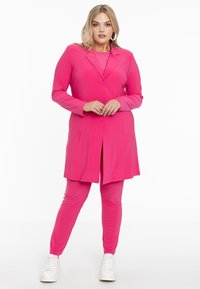 Felroze pak met een lange blazer en strakke broek. De blazer heeft een revers en drie knopen. G gedragen met witte sneakers. Glad stof.