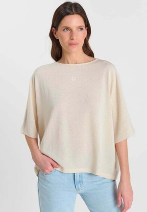 Vrouw met bruin haar, gekleed in een losse, lichtbeige trui met korte mouwen en lichtblauwe spijkerbroek, staand met één hand in de zak.