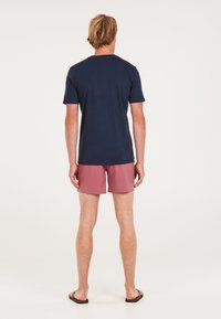 Camiseta de manga corta azul marino con cuello redondo, combinada con pantalones cortos rosas, vista desde la parte posterior. Se llevan sandalias planas en los pies.