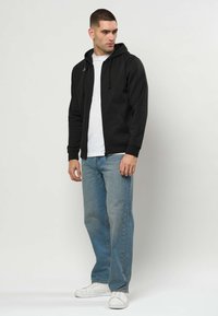 Melnā rāvējslēdzēja kapuce ar aukliņām, valkāta pār baltu t-kreklu, kopā ar gaiši ziliem džinsiem un baltiem sporta apaviem. Vienkāršs, ikdienišķs dizains.