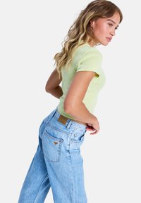 Jasnozielony krótki top w połączeniu z jasnoniebieskimi jeansami z wysokim stanem. Jeansy mają metkę z logo oraz wzór serca na tylnej kieszeni.