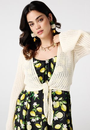 Une femme aux longs cheveux noirs porte un cardigan en tricot crème et une robe à motif citron, accessoirisée avec des boucles d'oreilles florales et un collier en chaîne.
