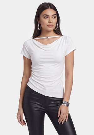 Jonge vrouw met lang donker haar draagt een wit topje met korte mouwen en een unieke halslijn, met zwarte leren broek, geaccentueerd met grote ringoorbellen en een zilveren manchet.