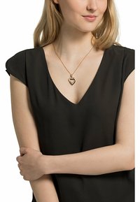 Guldtonet hjerteformet vedhængs halskæde med en indre sølvaccent, båret på en sort v-hals bluse. Enkel kæde design.