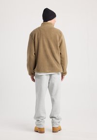 Giacca in pile marrone con colletto alto, abbinata a pantaloni felpati grigi chiari. Il modello indossa anche un berretto nero e stivali tan.