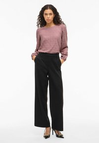 Sieviete ar viļņainiem matiem, uzvilkusi mauve krāsas teksturētu garām piedurknēm blūzi, melnus platības bikses un melnas papēžu kurpes ar zelta ķēdes detaļu.