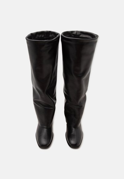 Paire de bottes hautes en cuir noir avec des tiges légèrement amples et des bouts carrés, vues de dessus sur fond blanc.
