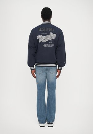 Mørkeblå bomberjakke med ribbede manchetter og kant, prydet med hvid broderet tekst på ryggen. Sammen med lyseblå flare-jeans og sneakers.