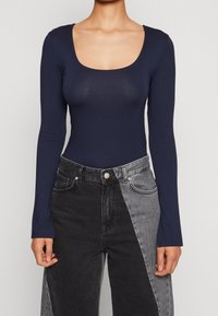 Marinblå långärmad bodysuit med rundad halsringning, tillverkad av ett mjukt, stretchigt tyg. Matchad med hög midja, tvåtonade svarta och grå jeans.