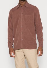 Man wearing a long-sleeve button-up shirt in muted mauve with a chest pocket, paired with beige pants against a plain background.