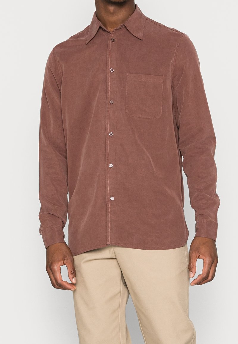 Man wearing a long-sleeve button-up shirt in muted mauve with a chest pocket, paired with beige pants against a plain background.