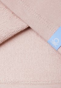 Tessuto rosa tenue con trama morbida, bordo a maglia fine e un'etichetta azzurro chiaro con un piccolo logo bianco, applicata all'angolo.