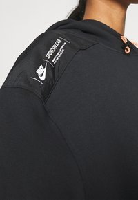 Svart hoodie med texturerad tyg, utrustad med en glänsande axelpanel som visar vita logotyper och en orange dragsko.