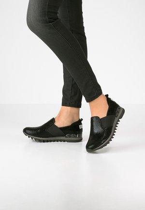 Person iført sorte teksturerede slip-on sko med tykke, rillede såler og sorte tilpassede bukser, stående på en hvid gulv.