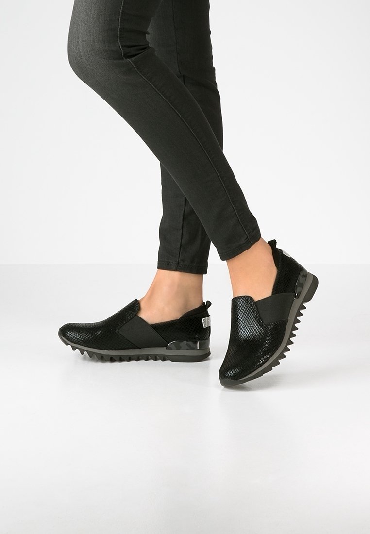 Personne portant des chaussures noires sans lacets à texture, avec des semelles épaisses et rainurées, et un pantalon moulant noir, debout sur un sol blanc.