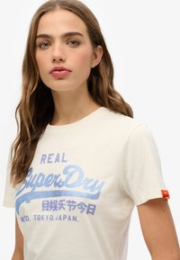 T-shirt couleur crème avec un texte graphique bleu "REAL SuperDry" et des caractères japonais. Manches courtes et une étiquette logo orange sur la manche.