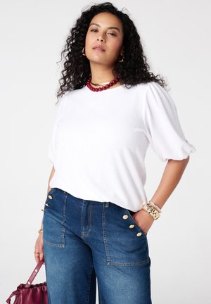 Vrouw met krullend haar die een wit topje met pofmouwen draagt, een high-waist blauwe spijkerbroek met gouden knopen, een rode kralenketting en een bordeauxrode handtas vasthoudt.