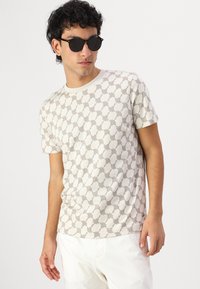 T-shirt à manches courtes, couleur crème, avec un motif géométrique marron. Tissu doux avec un col rond. Porté avec des lunettes de soleil foncées et un pantalon clair.