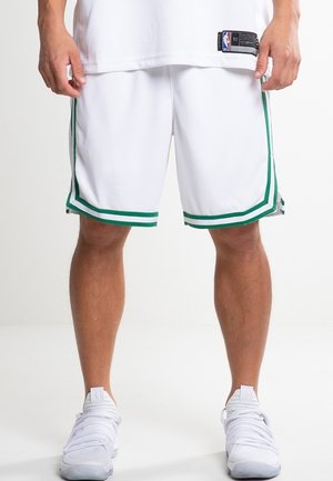 Homme portant un short de basketball blanc avec des bandes vertes et des chaussures de sport blanches, debout devant un fond clair uni.