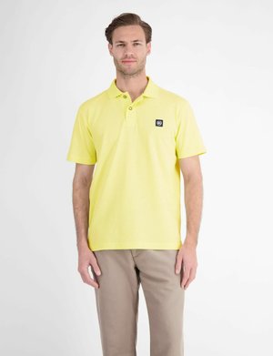 Mand står iført en lysegul kortærmet polo shirt med et lille sort logo mærke og beige bukser mod en hvid baggrund.