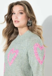 Młoda kobieta o falowanych jasnobrązowych włosach ma na sobie jasnozielony sweter z różowymi sercami oraz duże metaliczne kolczyki w kształcie kwiatów.