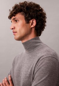 Joven con cabello castaño rizado, que lleva un jersey de cuello alto gris, mirando hacia la izquierda sobre un fondo liso.