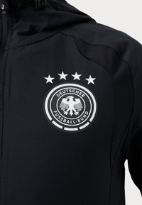 Musta urheilutakki, jossa on suuri, pyöreä valkoinen läiskä, jossa on kaksipäinen kotka -logo ja viisi tähteä sen yläpuolella.