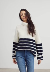 Suéter crema con franjas horizontales en azul marino, con cuello alto y hombros caídos. Combinado con jeans azules. Textura de punto suave.