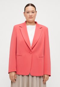 Korallfarget blazer i et glatt stoff, med én knapp, klaffelapper og frontlommer, brukt over en hvit topp og en plissert beige skjørt.