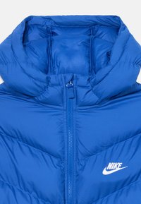 Mėlyna pūkinė striukė su siuvinėta medžiaga, aukštu kaklu, priekiniais užtrauktukais ir balta Nike logotipu kairėje pusėje. Lygus, blizgus audinys.