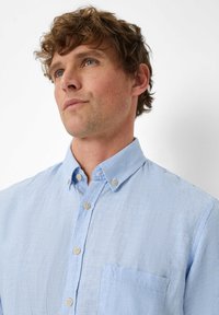 Camicia a quadri azzurro chiaro con colletto button-down, taschino sul petto e bottoni beige. Presenta un tessuto testurizzato con un motivo sottile.