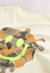Cream-colored t-shirt featuring a camouflage pattern in brown and orange, with a large green smiley face graphic in the center.