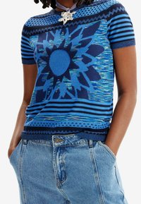 Jersey azul de manga corta de punto con un diseño de girasol, rayas texturizadas y un borde de encaje en el dobladillo, combinado con jeans de mezclilla azules.