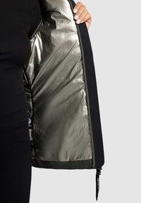 Schwarze Jacke mit metallisch silbernem Futter, Reißverschluss und kontrastierenden grünen Akzenten. Hat eine glatte Textur und ein Etikett im Inneren.